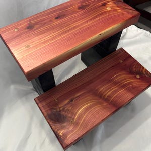 May include: Two-tiered wooden step stool with a rich, reddish-brown finish. The steps are rectangular, showcasing the wood grain and knots. The supports are painted black, creating a contrast. A handcrafted item.