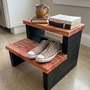 May include: A two-step wooden stool featuring natural reddish-brown wood grain treads and solid black side supports. The top step holds a brown ceramic mug and two stacked books, while the bottom step holds a pair of gray canvas sneakers.