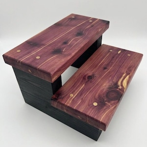 May include: A two-step stool featuring natural reddish-purple cedar wood treads with visible knots and grain patterns. The steps are secured with small circular brass-colored fasteners, and the base structure is finished in a solid matte black.
