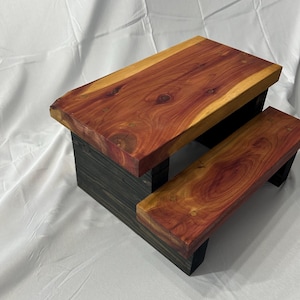 May include: A small wooden step stool with a natural wood top and a dark stained base. The top is made of reddish-brown wood with visible grain patterns. The base is a dark gray color. The stool has a simple, functional design.