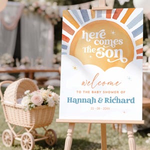Può includere: Un cartello di benvenuto rettangolare su un cavalletto in legno con un grafico solare ad acquerello con raggi blu e arancioni. Il testo recita: here comes the Son, welcome TO THE BABY SHOWER OF Hannah & Richard, e 22 - 08 - 20XX, con piccole stelle.