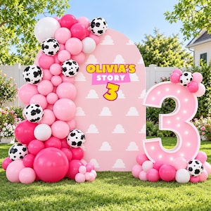 Könnte beinhalten: Ein rosafarbener, bogenförmiger Hintergrund mit weißem Wolkenmuster und dem Text OLIVIA'S STORY 3, verziert mit einer Ballongirlande in Rosa, Weiß und Kuhfleckenmuster. Rechts steht eine große, beleuchtete rosa Zahl 3 mit passenden Ballons.