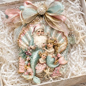 May include: A decorative ornament featuring a Santa Claus merman and two mermaid figures centered within a large scalloped seashell. The piece is adorned with pastel pink and blue ribbons, a pearl accent, and small starfish, all nestled in a gift box.