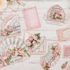 May include: A collection of paper ephemera featuring pink roses, lace patterns, and vintage-style text including "ENVI DE PARIS" and "RICHARDS BROTHERS". The set includes a lined index card, a postcard, a ticket, and various decorative die-cut shapes.