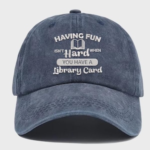 Peut inclure: Casquette de baseball bleu marine avec l'inscription blanche "HAVING FUN ISN'T HARD WHEN YOU HAVE A library Card". La casquette a une visière incurvée et un bouton sur le dessus. Le texte comprend une image d'un livre ouvert.