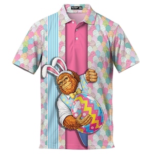 May include: A short-sleeved polo shirt with a colorful Easter egg pattern and a pink center panel. The shirt features a cartoon Sasquatch wearing bunny ears and holding a decorated egg. The shirt is a size 3XL.