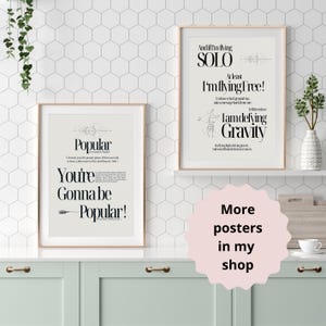 Op de afbeelding: Twee afdrukbare posters met zwarte tekst op een witte achtergrond. De eerste poster zegt "Popular You're Gonna Be Popular!" De tweede poster zegt "And if I'm flying SOLO At least I'm flying free! I am defying Gravity". De posters zijn ingelijst en hangen aan een witte muur met een zeshoekig patroon.