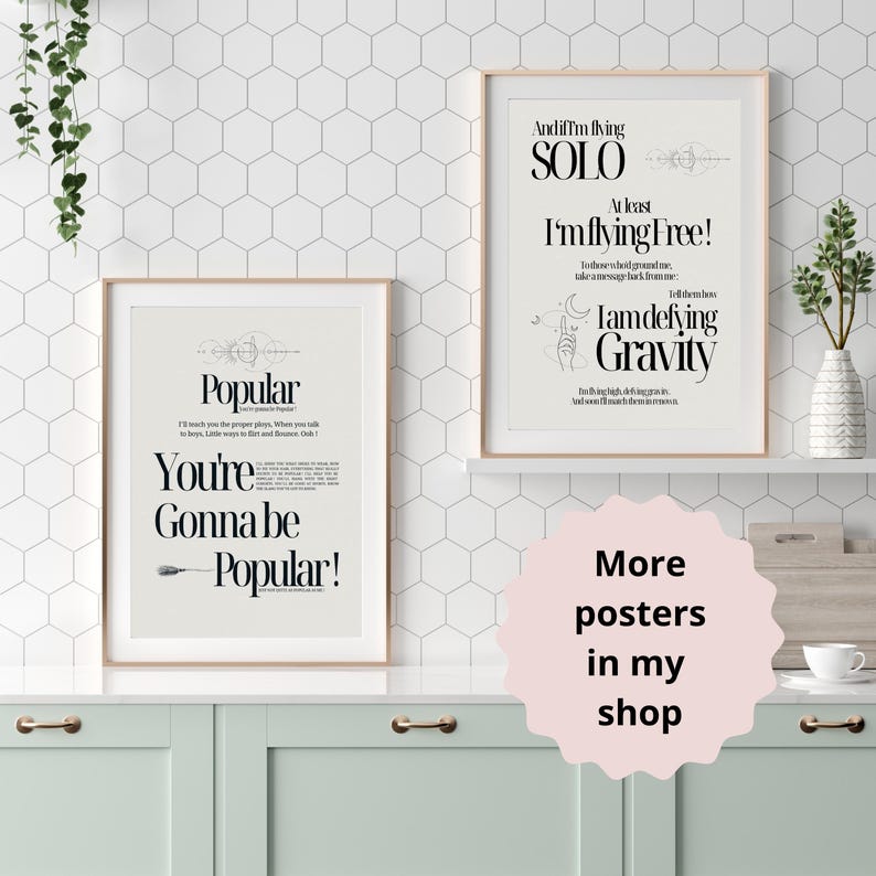 Op de afbeelding: Twee afdrukbare posters met zwarte tekst op een witte achtergrond. De eerste poster zegt "Popular You're Gonna Be Popular!" De tweede poster zegt "And if I'm flying SOLO At least I'm flying free! I am defying Gravity". De posters zijn ingelijst en hangen aan een witte muur met een zeshoekig patroon.