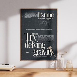 Puede incluir: Una impresión artística enmarcada con las palabras "It's time to try defying gravity" en blanco sobre un fondo oscuro. La impresión incluye texto adicional y elementos decorativos, exhibidos sobre una superficie de madera.