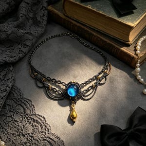 May include: A dark grey macrame necklace featuring a central circular blue cabochon stone. The design includes intricate woven cord patterns, small gold-toned metallic beads, and a single gold-toned teardrop-shaped pendant hanging from the centre.
