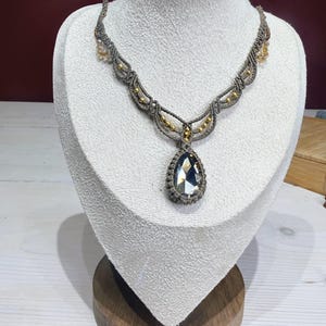 May include: A macrame necklace featuring a large, faceted, teardrop-shaped gray crystal pendant centered on a knotted gray cord. The necklace is embellished with small, metallic gold-toned beads integrated into the intricate macrame pattern.