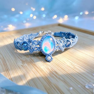 May include: A light blue macrame bracelet featuring a central oval cabochon with swirling pink and blue patterns. The band is woven with blue cord, accented by small white pearls, silver beads, and two faceted dark blue crystal beads on each side.