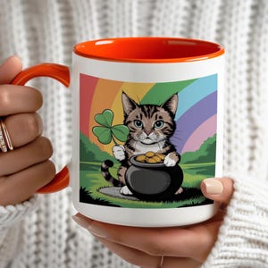 May include: A white ceramic mug with an orange rim features a cartoon cat holding a pot of gold and a four-leaf clover. The cat is in front of a rainbow.