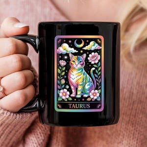 May include: Black ceramic mug with a vibrant illustration of a cat with rainbow fur, flowers, clouds, and a crescent moon. The word "TAURUS" is at the bottom. The mug is held by a person.