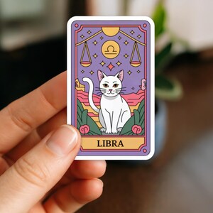 May include: A sticker featuring a white cat illustration within a purple and gold frame. The design includes Libra scales, stars, and the word "LIBRA" at the bottom. The cat is sitting in front of a landscape with pink and green elements.