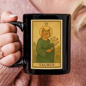 May include: Black ceramic mug featuring a vintage-style illustration of a cat dressed in green robes, holding flowers, and a gold halo. The image is a tarot card design with the word "TAURUS" at the bottom.