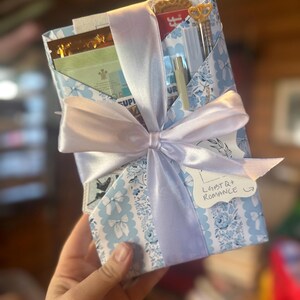 以下が含まれることがあります： 水色の花柄の紙で包まれ、幅広の淡い紫色のサテンリボンで結ばれたギフトボックス。 クッキー、ペン、小さなボトルなど、さまざまなアイテムが入っています。 タグには「LGBTQ+ Romance」と書かれています。