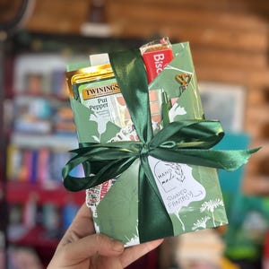 以下が含まれることがあります： 緑色のギフトボックス。花柄で、濃い緑色のリボンで結ばれています。中にはお茶、ビスケット、小さなギフトが入っています。「Handmade Signed Fantasy」と書かれたタグが付いています。