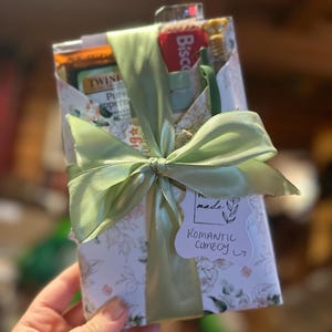 以下が含まれることがあります： 花柄の紙で包まれ、明るい緑のリボンで結ばれたギフトボックス。箱には、お茶、ビスケット、歯ブラシなど、さまざまなアイテムが入っています。小さなタグには「Romantic Comedy」と書かれています。