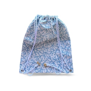 Puede incluir: Una mochila con cordón con un fondo blanco y un patrón repetitivo de coral azul. La bolsa tiene cordones blancos y ojales y topes de cordón de metal dorado. La bolsa está hecha de una tela ligera.
