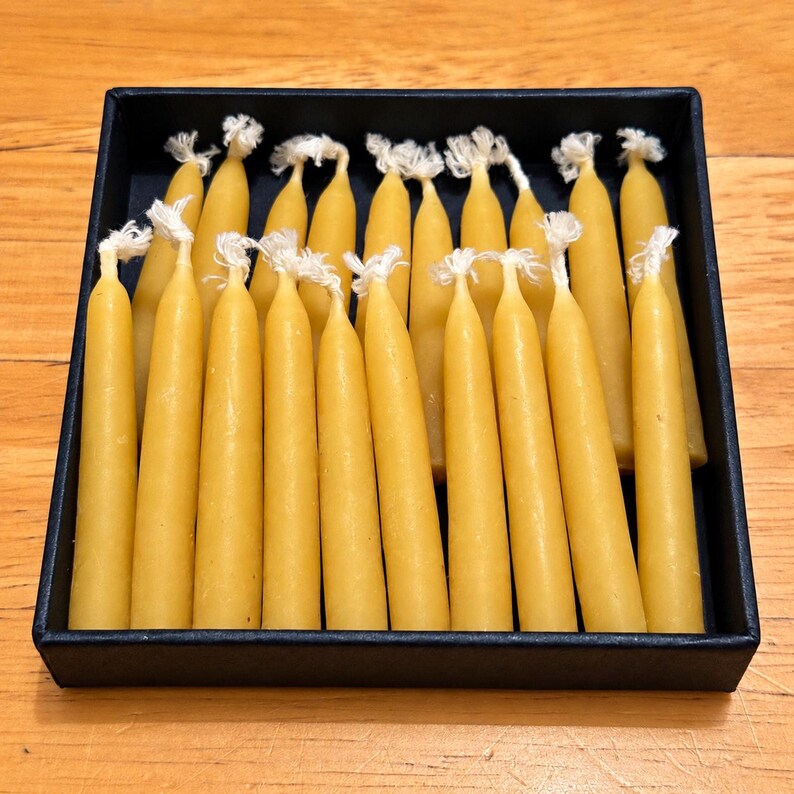 May include: A collection of thin, cylindrical yellow beeswax candles with visible white cotton wicks, arranged upright in a square black box. The candles are uniform in color and shape, filling the container in a dense, organized grid formation.