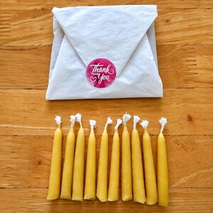 May include: Ten yellow beeswax taper candles with white cotton wicks arranged in a row. Above the candles sits a white paper package sealed with a round pink sticker featuring the white text Thank You and a small heart icon.