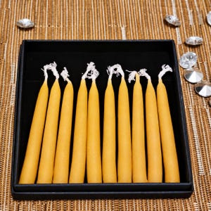 May include: A set of ten slender, golden-yellow taper candles arranged vertically in a black rectangular box. Each candle features a smooth, cylindrical body with a visible white cotton wick at the top, presented in a uniform row.