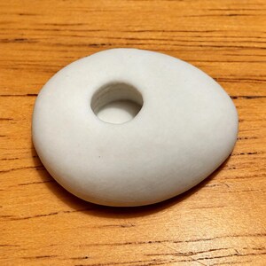 May include: A smooth, matte white ceramic object with an asymmetrical, pebble-like shape. The piece features a single, circular recessed opening in the center, creating a small, hollow basin within the solid, rounded form of the white ceramic item.