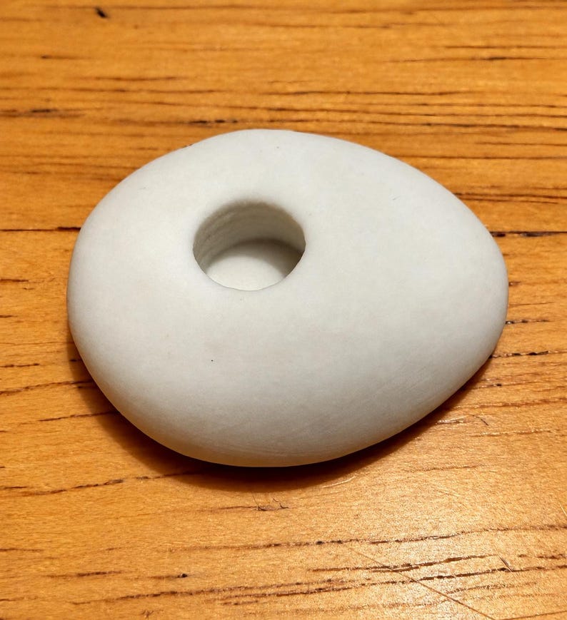 May include: A smooth, matte white ceramic object with an asymmetrical, pebble-like shape. The piece features a single, circular recessed opening in the center, creating a small, hollow basin within the solid, rounded form of the white ceramic item.