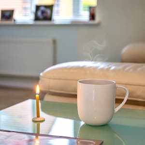 May include: A plain white ceramic mug with a curved handle sits next to a thin yellow taper candle standing in a small white circular base. The mug emits a faint wisp of steam, indicating a hot beverage inside, while the candle flame glows.