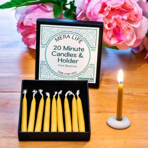 May include: A black box containing eight yellow beeswax candles, with a label reading MERA LIFE 20 Minute Candles & Holder Pure Beeswax Time For You. A single lit yellow candle stands in a small gray circular holder next to the box.