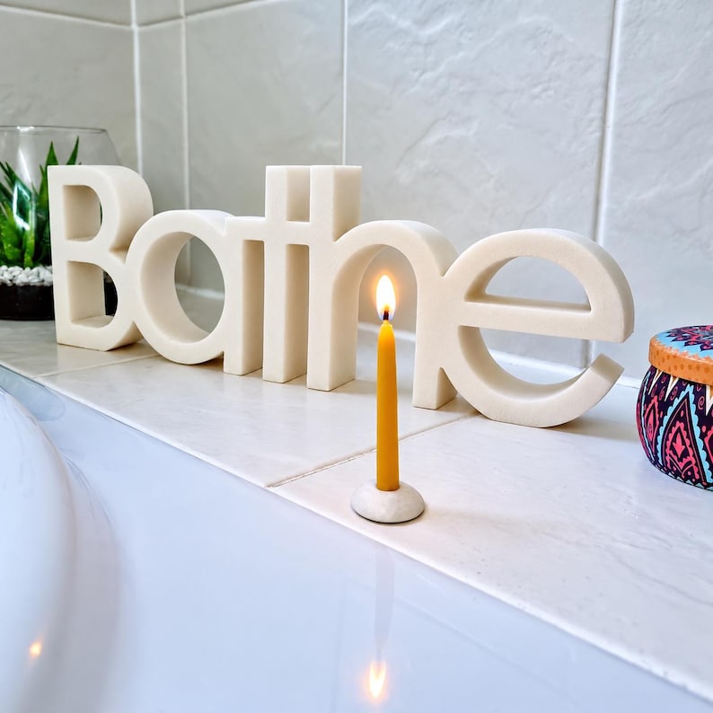 May include: A cream-colored, three-dimensional sign spelling the word Bathe in a rounded, sans-serif font. In front of the sign sits a thin, yellow beeswax candle with a lit flame, held in a small, circular, light-colored stone candle holder.