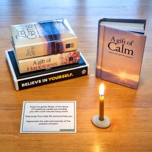 May include: A collection of four books titled Zen Gems, A Gift of Happiness, Believe in Yourself, and A Gift of Calm, alongside a small lit beeswax candle in a white holder and a rectangular card with printed text about enjoying the candle flame.