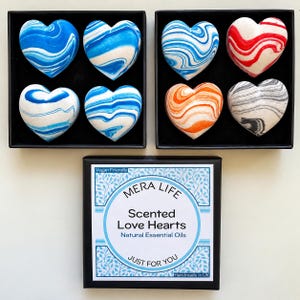 May include: Two black boxes containing four heart-shaped items each, featuring blue, white, red, orange, and grey marbled patterns. A third black box features a label reading MERA LIFE Scented Love Hearts Natural Essential Oils Vegan Friendly.