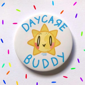 May include: A white button with a blue border and the text "DAYCARE BUDDY" in blue. A yellow sun with a smiling face and pink cheeks is in the center of the button.
