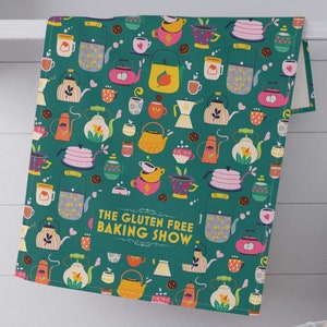 May include: A green tea towel with a colorful pattern of teapots, cups, and flowers. The text "THE GLUTEN FREE BAKING SHOW." is printed on the towel.