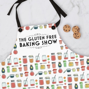 Könnte beinhalten: Weisse Schürze mit schwarzem Nackenband und dem Text "THE GLUTEN FREE BAKING SHOW". Die Schürze hat ein Muster aus verschiedenen Gläsern und Keksen. Die Schürze ist zum Backen und Kochen bestimmt.