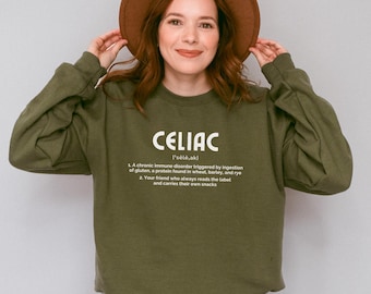 May include: An olive green sweatshirt featuring the word "CELIAC" in white, with a definition below. The sweatshirt is worn by a person wearing a brown hat. The definition describes a chronic immune disorder triggered by gluten.