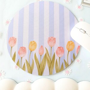 Op de afbeelding: Ronde muismat met een bloemenontwerp. Het heeft een lichtblauwe en witte gestreepte achtergrond met aquarel tulpen in roze en geel. Een witte computermuis is zichtbaar aan de rechterkant.