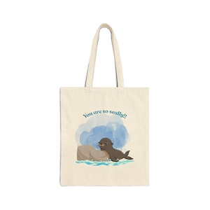 May include: Cream-colored tote bag featuring a cartoon seal illustration. The seal is brown and black, resting on a rock with blue water around it. The text "You are so seally!!" is above the seal in blue.