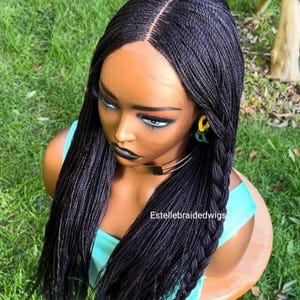 May include: A black braided wig with a lace front. The wig is styled in long, straight braids with two braids on each side of the face. The wig is on a mannequin head and is set against a green grassy background.