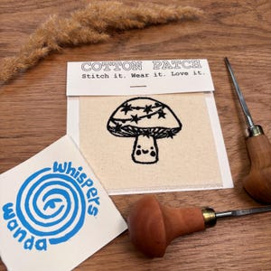 Op de afbeelding: Een beige katoenen patch met een zwart paddestoelontwerp en de tekst "COTTON PATCH Stitch it. Wear it. Love it." Een blauw-wit logopatch met de tekst "whispers wanda" is ook zichtbaar. Een houtsnijgereedschap staat op de achtergrond.