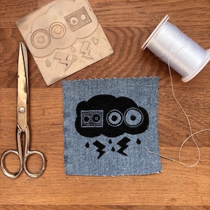 Könnte beinhalten: Eine Schwarzweißillustration einer Wolke mit einem Ghettoblaster und zwei Lautsprechern darin. Die Wolke regnet und es blitzt aus ihr heraus. Die Illustration ist auf einem blauen Jeans-Patch gedruckt.