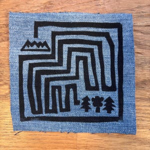 Peut inclure: Un motif noir et blanc sur un fond de denim bleu. Le motif présente un motif de labyrinthe avec une chaîne de montagnes en haut et une rangée d'arbres en bas.