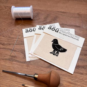 May include: A craft image featuring a spool of white thread, a needle, and a set of fabric patches. Each patch has a black bird design and the text "äöü". The text "FEATURING whispersofwanda" is also visible.