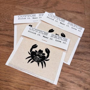Peut inclure: Trois écussons en coton crème avec un motif de crabe noir portant un chapeau et des lunettes. Chaque écusson est rectangulaire avec un fond crème et une bordure blanche. Le texte "COTTON PATCH Stitch it. Wear it. Love it." est imprimé sur une étiquette blanche.
