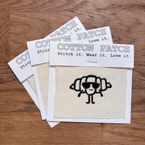 Könnte beinhalten: Drei Baumwollflicken mit einem schwarzen, gestickten Design eines Croissants mit Sonnenbrille. Jeder Flicken ist an einer weißen Karte mit dem Text "COTTON PATCH" und den Sätzen "Stitch it. Wear it. Love it." befestigt.