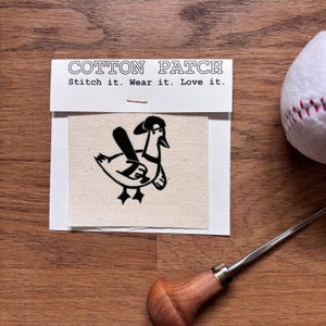 Könnte beinhalten: Ein Baumwollflicken mit dem Text "Cotton Patch" und "Stitch it. Wear it. Love it." Der Flicken zeigt eine schwarz-weiße Illustration einer Ente mit Baseballkappe und Baseballschläger. Ein Baseball und ein Werkzeug sind ebenfalls im Bild.