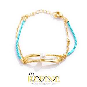 May include: A gold bracelet featuring a turquoise rope and a gold chain. The bracelet has a gold bar with a pearl in the centre. The bracelet is on a white background.