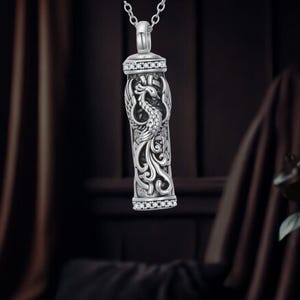Może przedstawiać: Srebrzysty, cylindryczny wisiorek na łańcuszku z wytłoczonym wzorem feniksa i misternymi zawijasami. Górna i dolna krawędź cylindra są ozdobione rzędem małych, przezroczystych i błyszczących kamieni osadzonych w metalowej oprawie.